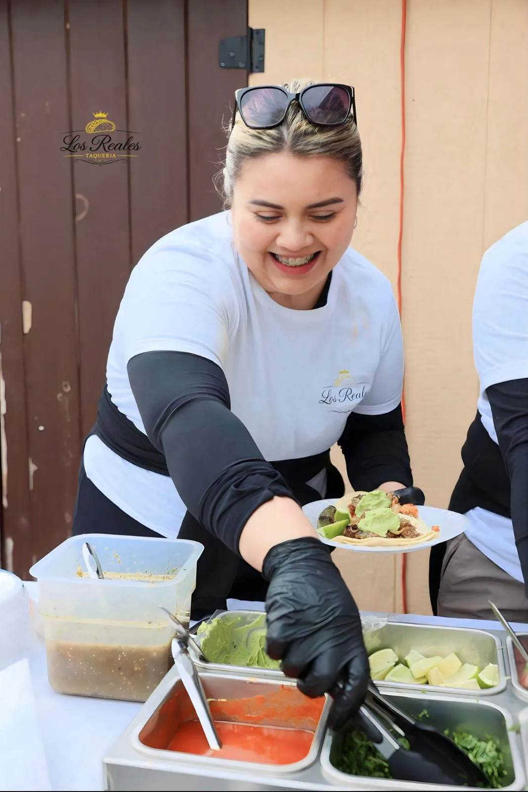 Integrante del equipo de Los Reales Taquería entrega un plato de tacos sobre la barra de salsa verde, salsa roja, guacamole y limones