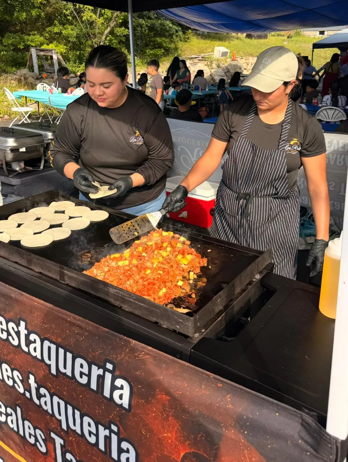 Dos taqueras de Los Reales Taquería cocinan carne adobada con piña en la plancha mientras las tortillas se calientan, en un evento al aire libre